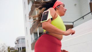 Woman running outside, wearing a phone armband. with Peloton app showing on the screen