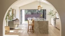 Large kitchen and dining area with curved archway, marble kitchen island, countertop stools, hanging lights, large plant in wicker pot, and large, open window revealing outdoor area.