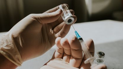 gloved hands draw fluid from a vaccine vial into a syringe