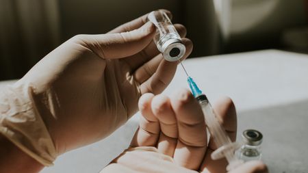 gloved hands draw fluid from a vaccine vial into a syringe