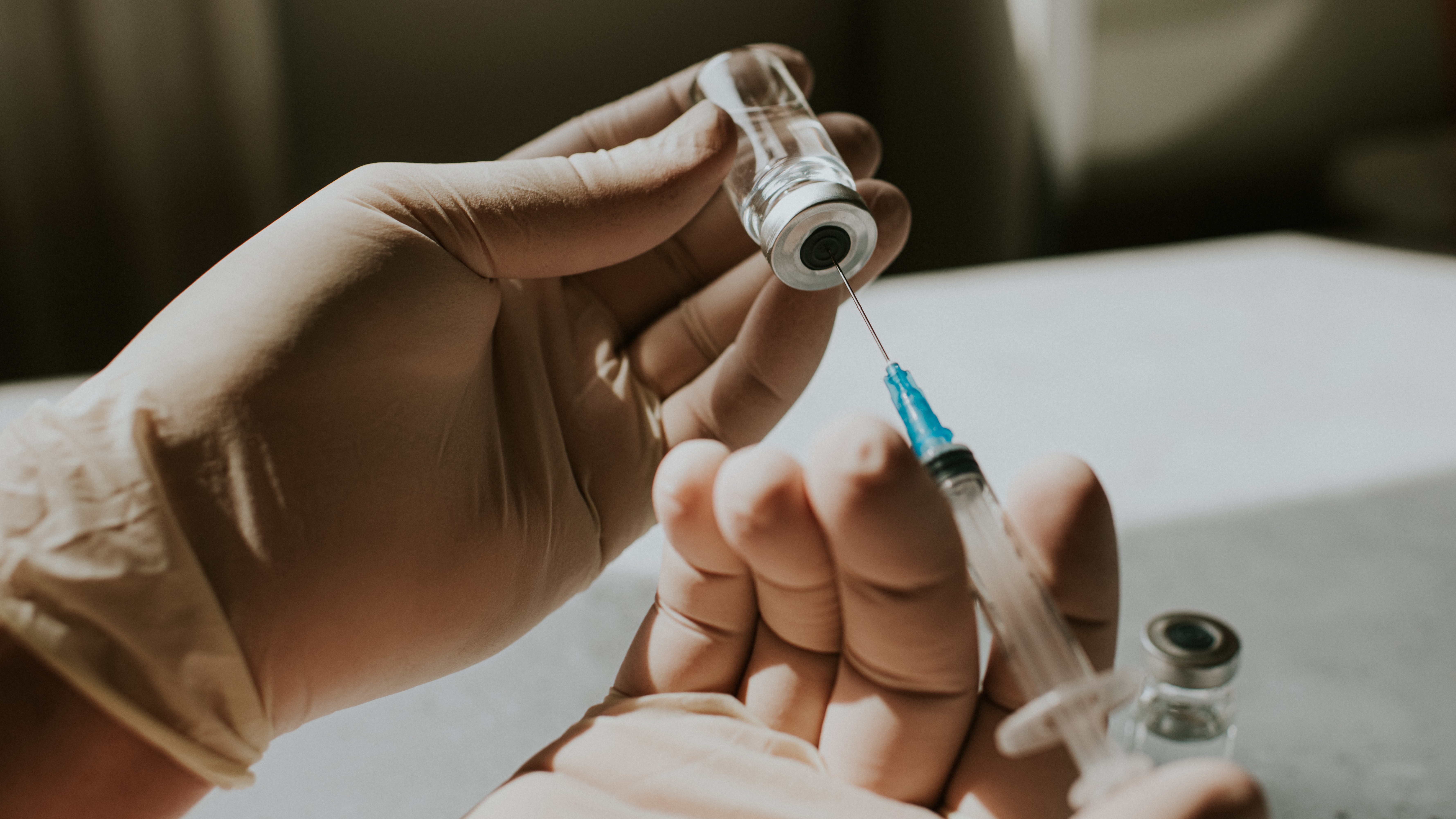 gloved hands draw fluid from a vaccine vial into a syringe