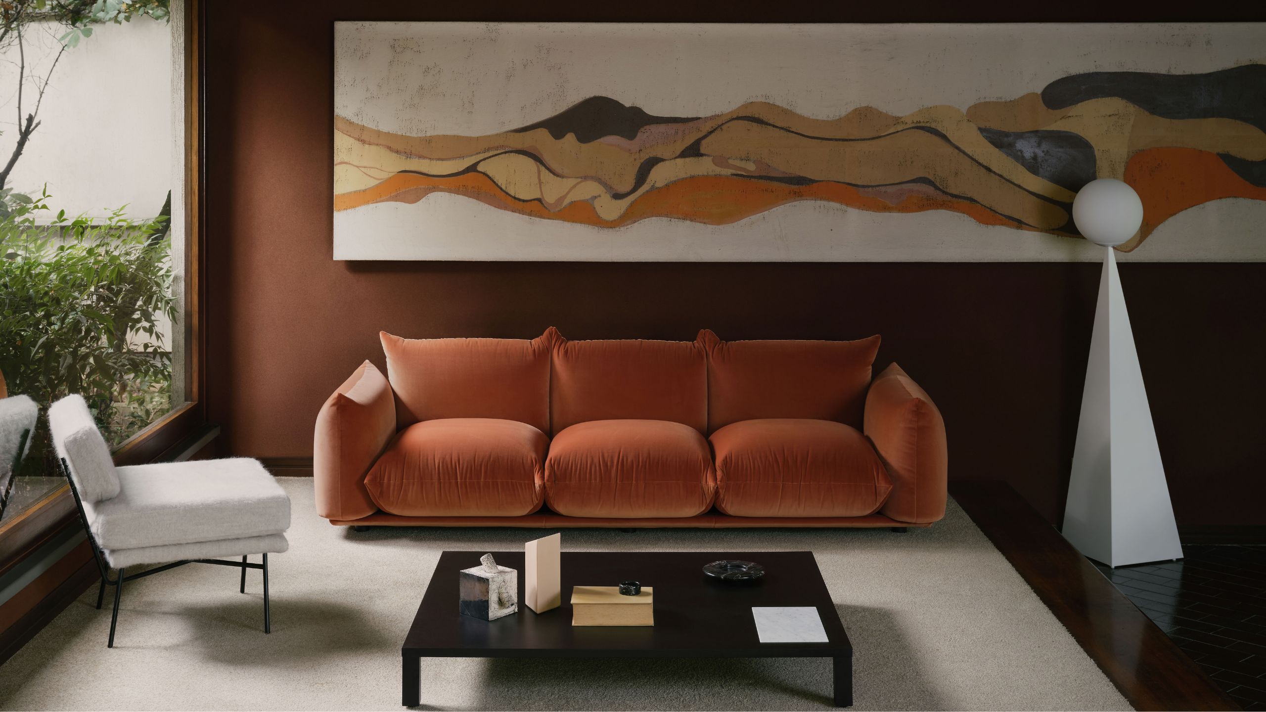 A modern living room with a bubble-style contemporary rust sofa, an abstract white floor lamp, and white accent chair, against a brown wall with a large artwork almost along the full length of it
