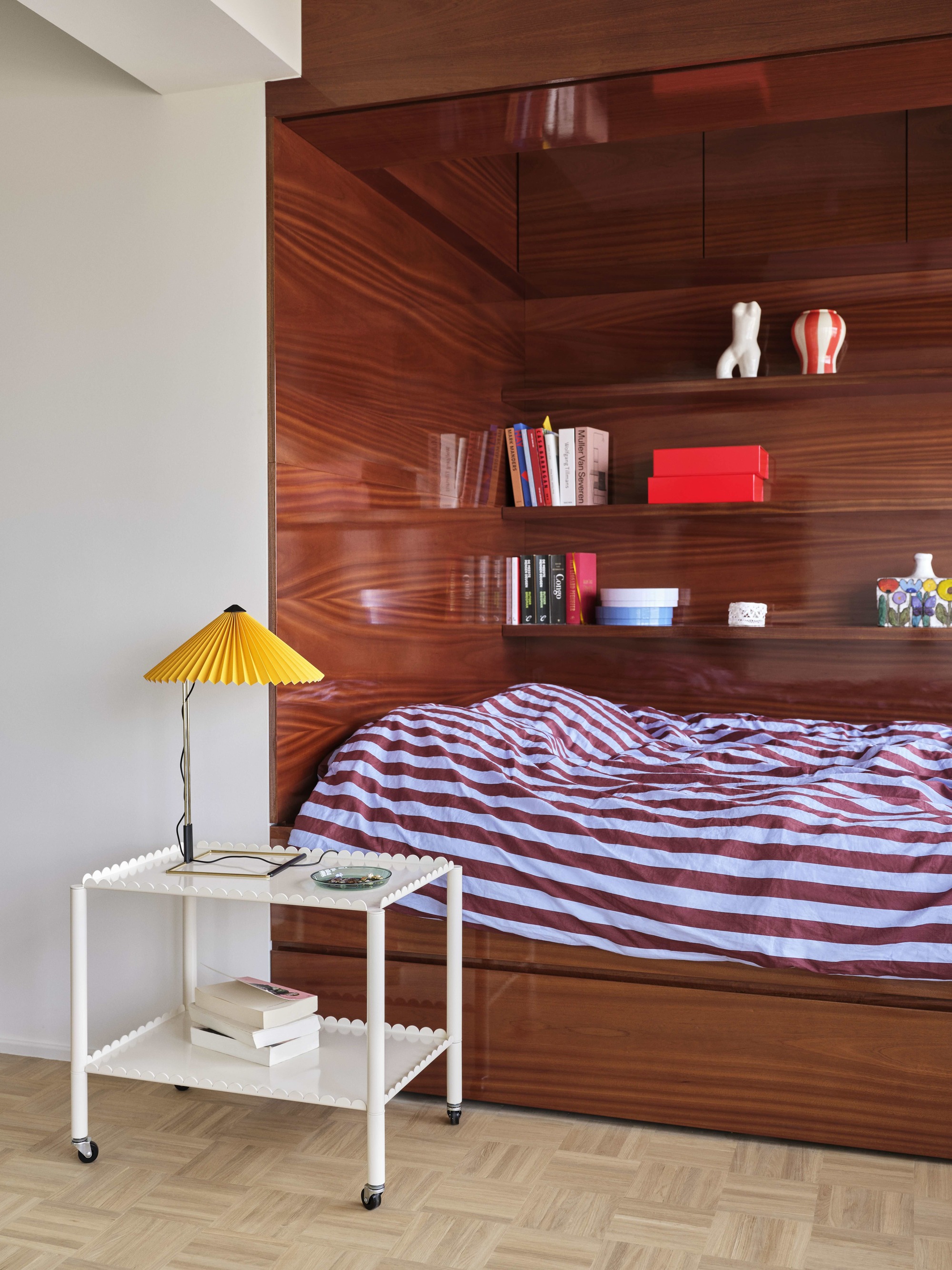 alcove bed with timber paneling, striped blue and brown duvet cover, floating wall shelves, a HAY scalloped side table and HAY lamp with yellow pleated lampshade