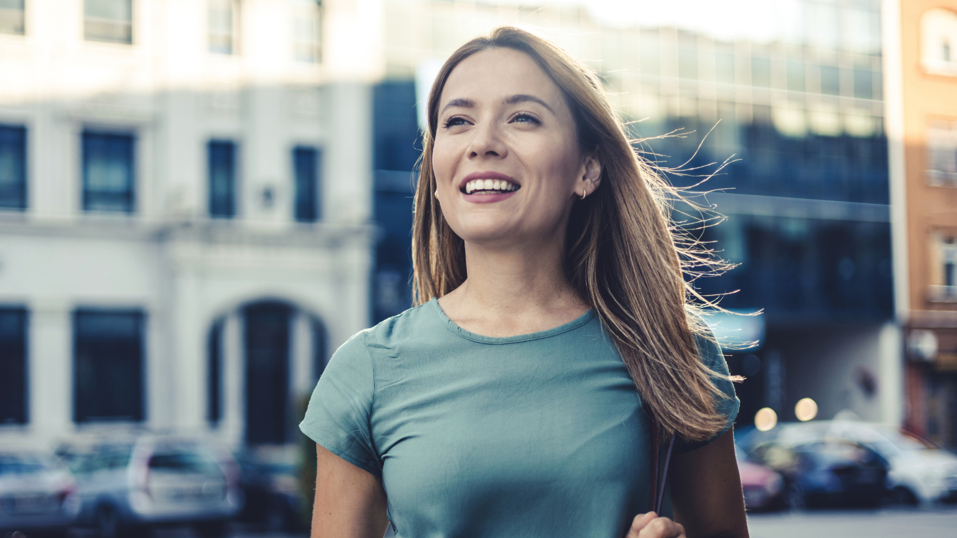 A woman wearing a teal t-shirt looking happy and energized as she walks along a street.