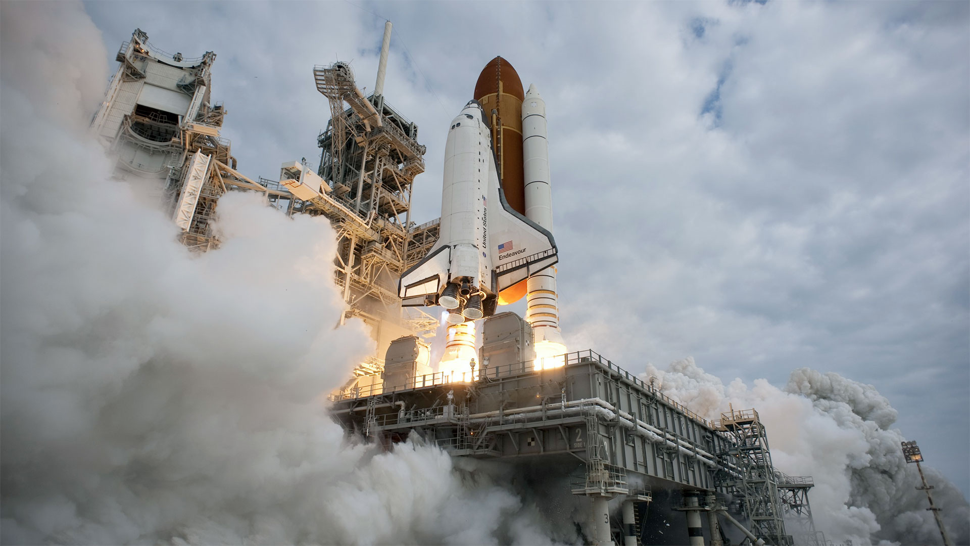 a black and white winged orbiter attached to a brownish-orange tank and two white boosters lifts off a metal-gray platform from the perspective of a camera position forward and underneath it.