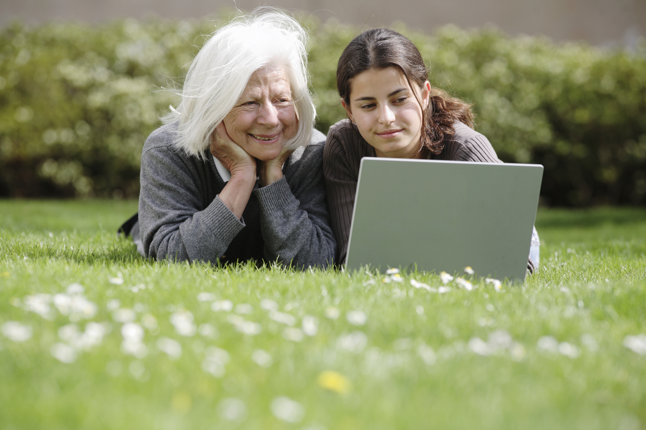 A grandmother and her granddaughter lie on the grass, looking at a laptop together.