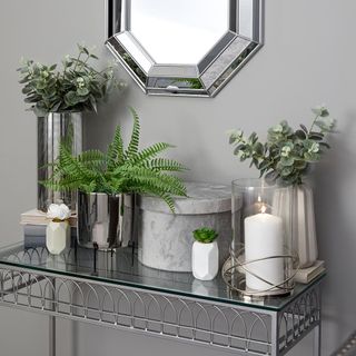Grey wall with silver mirror and a glass table with silver and white vases, green plants, and a storage box