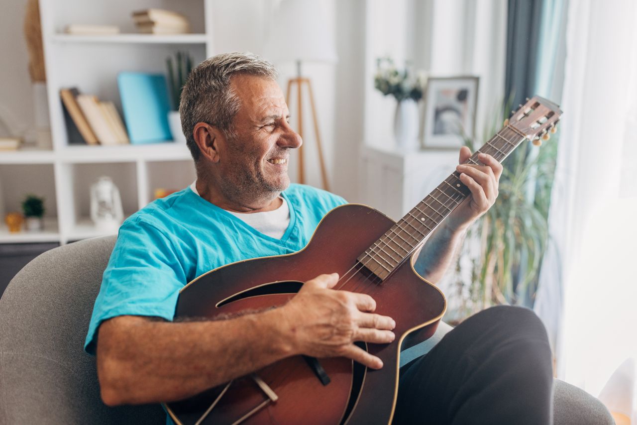 A smiling older man practices acoustic guitar at home.