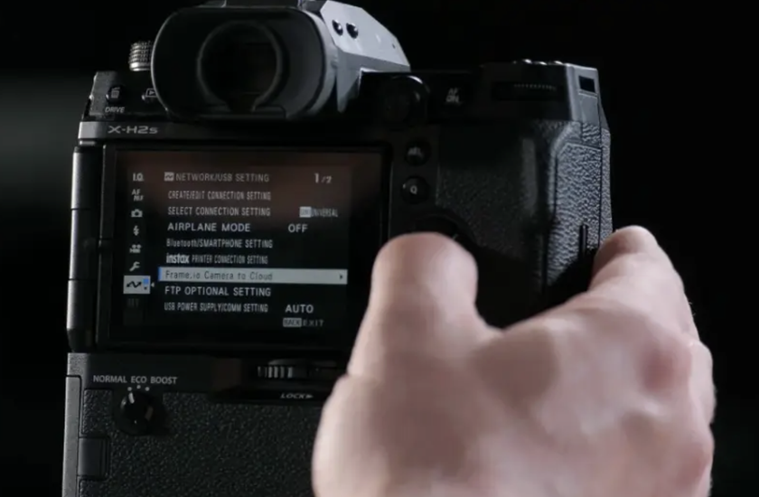 A close-up shot of the back of a black Fujifilm X-H2S camera shows the camera's LCD screen displaying the "NETWORK/USB SETTING" menu, with the option "Frame.io Camera to Cloud" highlighted and selected.