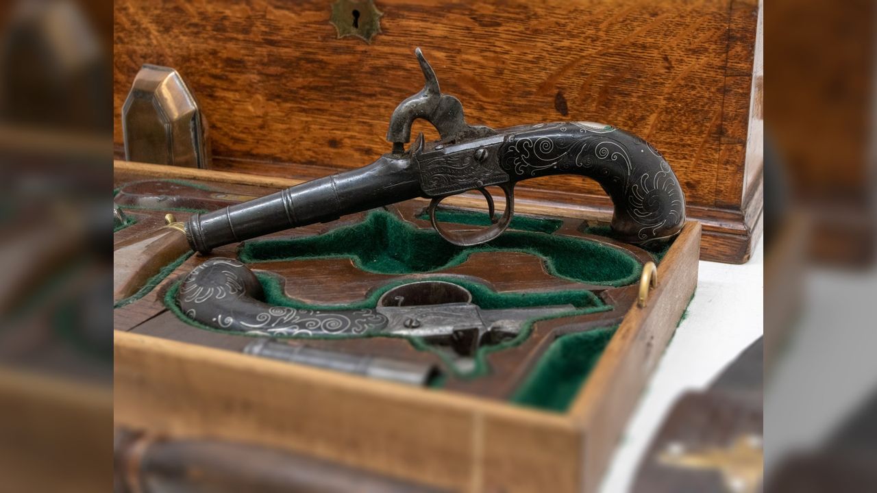 Here we see 2 old pistols in a special presentation case lined with green felt. The guns themselves have black handles with some white swirly patterns on them.