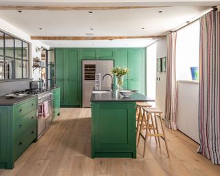 Modern kitchen in traditional property, bright green kitchen cabinet, kitchen island with wooden stools, light wooden flooring, white painted walls, beamed ceiling, window with long striped curtains