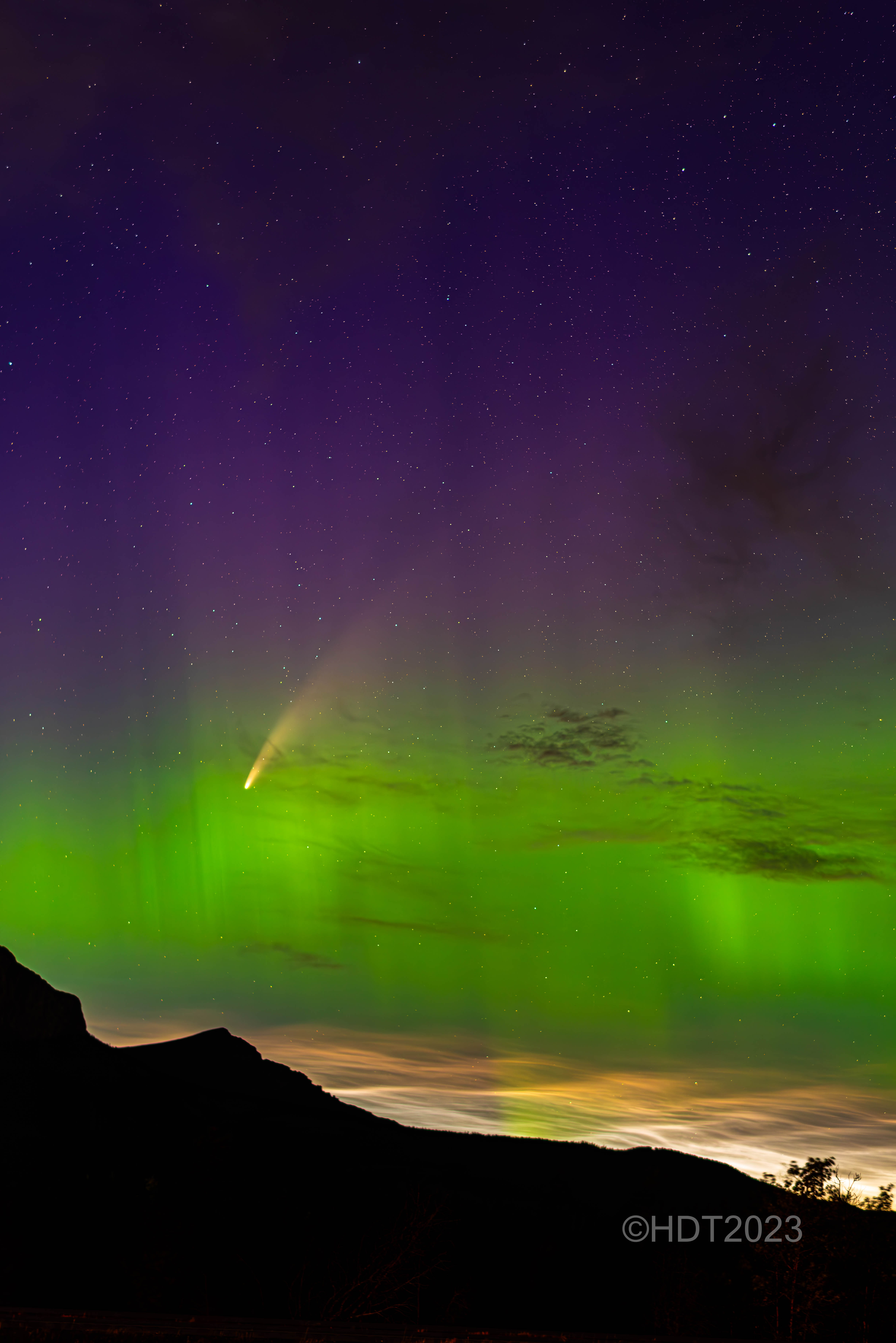 a bright comet streaks through curtains of purple and green light above thin wispy noctilucent clouds.