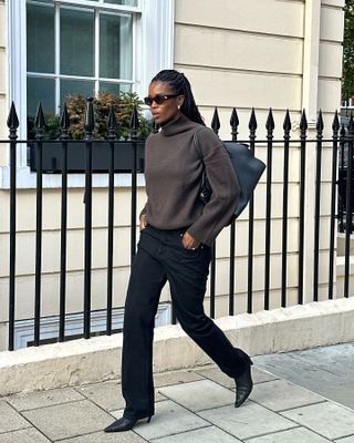 A woman wears a brown turtleneck sweater, black jeans, and black heeled booties.