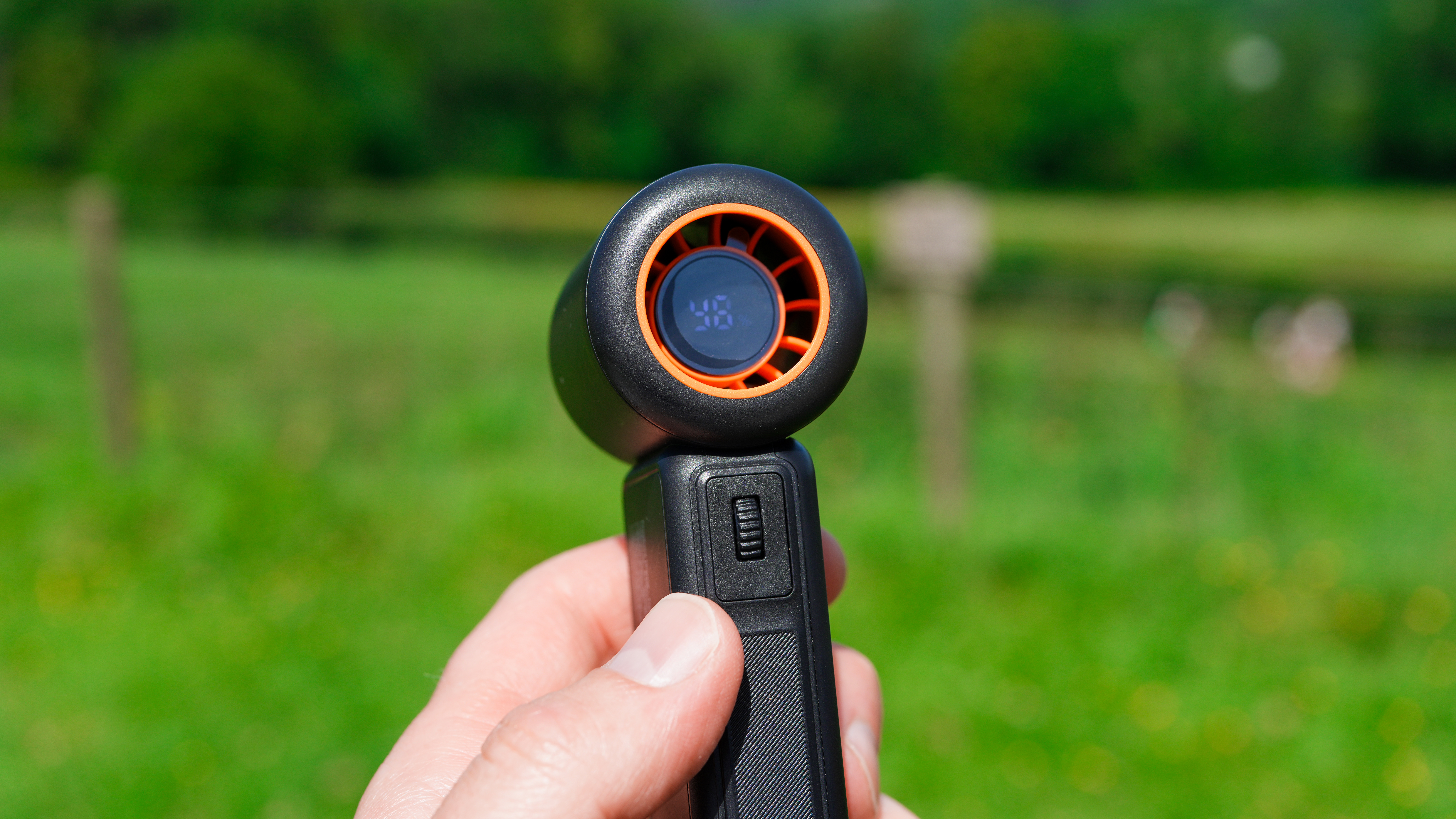 A Warmco portable handheld fan in hand with green foliage in the background