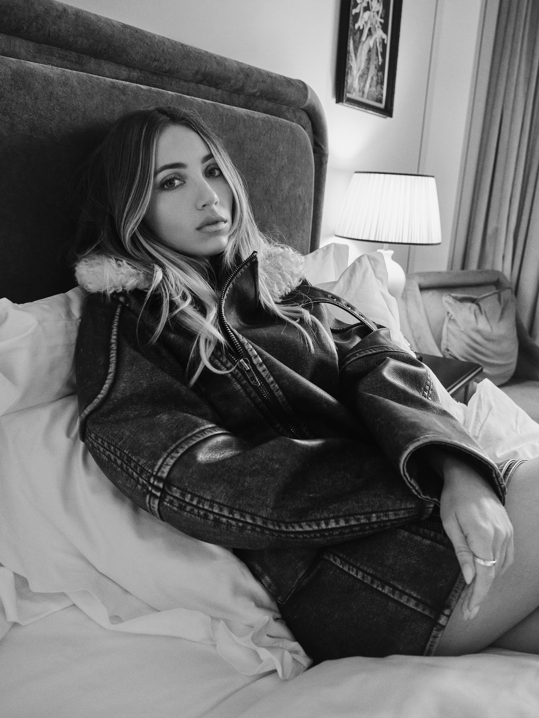 Emily Rudd photographed by Christian H&amp;ouml;gstedt. She is lying in bed in a hotel room wearing a leather jacket with fur collar.