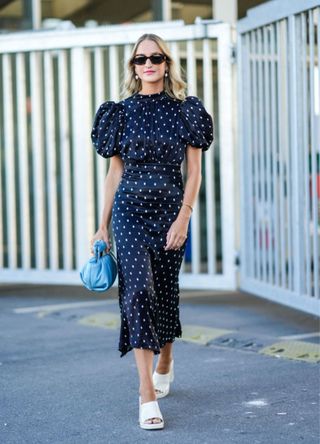 A guest wears black sunglasses, gold and pearl pendant earrings, a black with white polka dots print pattern short puffy sleeves / belted midi dress, a blue shiny leather puffy handbag from Karl Lagerfeld, gold rings, gold bracelets, white shiny varnished leather open toe-cap / block heels mules , outside Munthe during Copenhagen Fashion Week Spring/Summer 2023, on August 11, 2022 in Copenhagen, Denmark