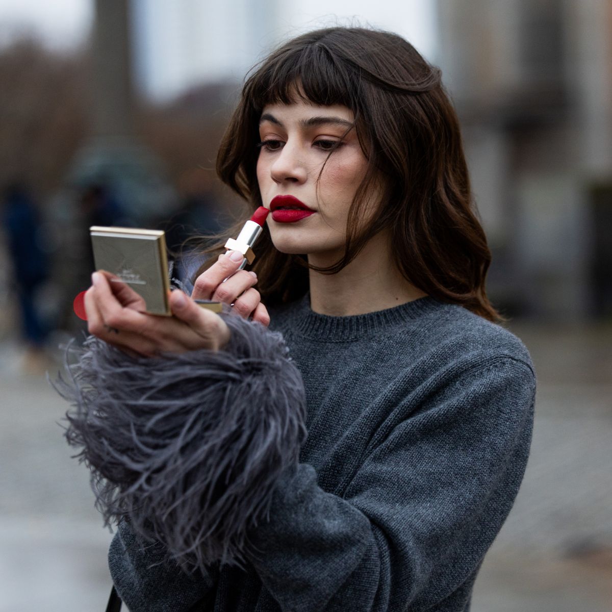street style shot of woman wearing grey jumper applying red lipstick