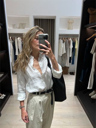 Eliza Huber in the dressing room at Banana Republic wearing a white linen button-down shirt with tan khaki pants, black slides, a black raffia oversize bag, and a black belt.
