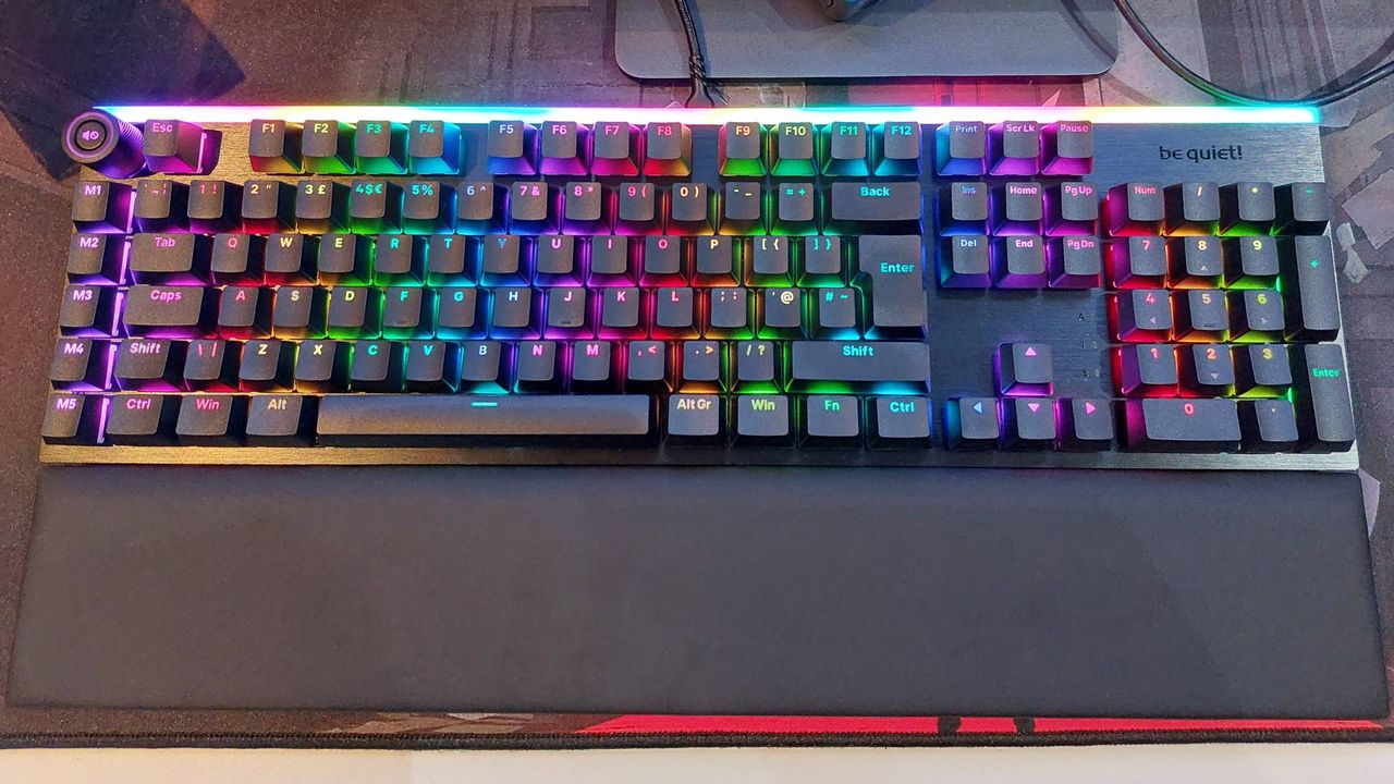Be Quiet's Light Mount mechanical gaming keyboard sits on a large red and black mouse mat. The magnetic wrist rest is attached. The RGB lights are on, displaying very bright and vibrant rainbow colours.