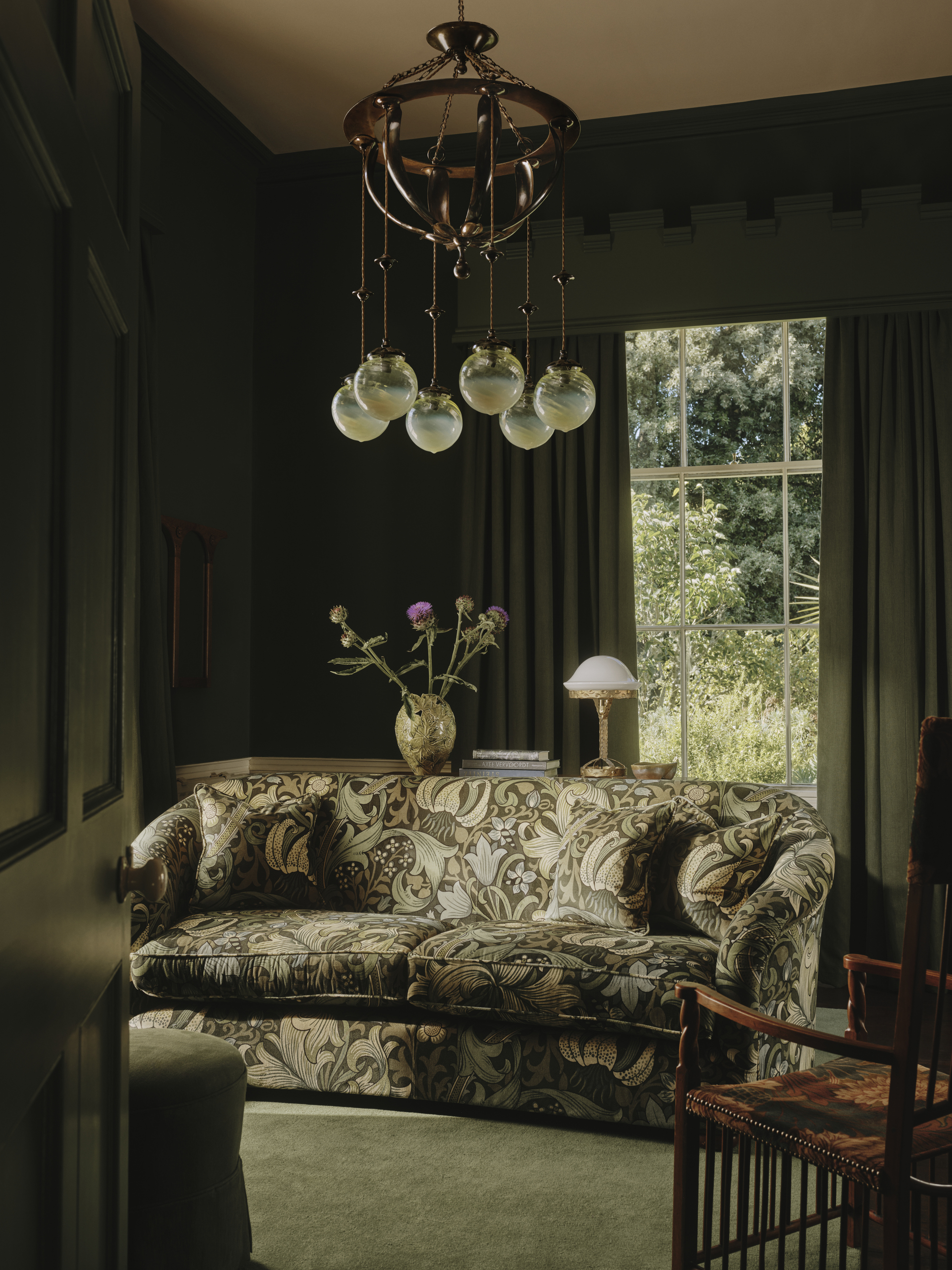 Image of a luxury living room with a green color palette. The walls are dark green, there is a light green carpet, and a green floral sofa with a pendant light overhead.
