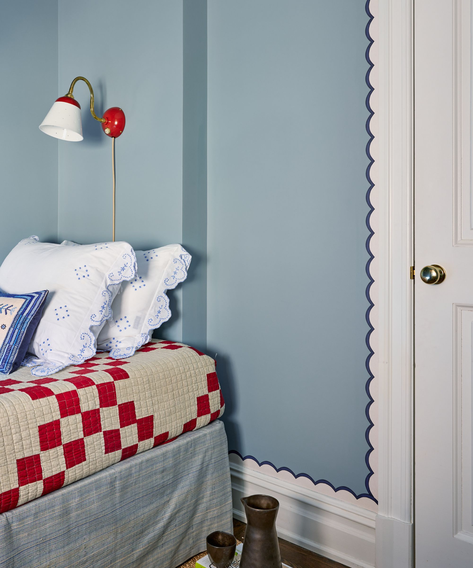 Corner of a bedroom with cornflower blue walls, a single bed with a red and white checkered quilt, and a doorway trimmed with a whimsical navy blue scalloped edge