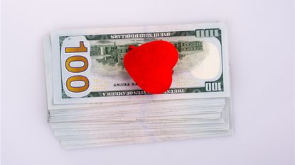 A heart knickknack sits on top of a stack of hundred-dollar bills.