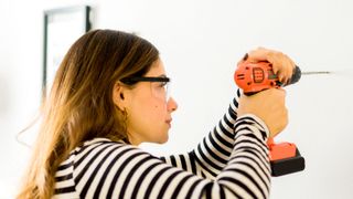 Brunette woman wearing black and white stripy top and safety glasses using an orange combi drill to drill into white wall