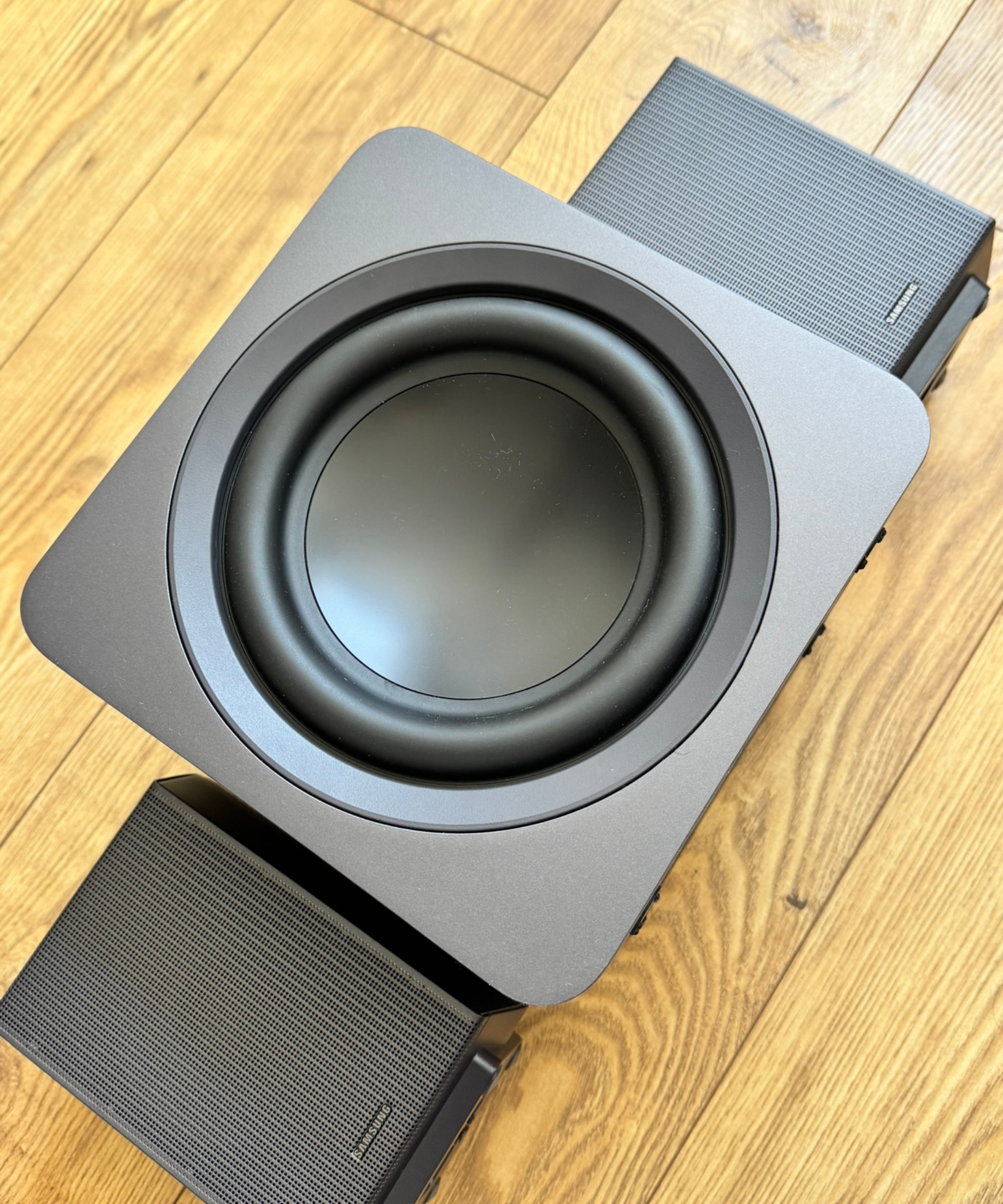 Samsung HW-Q990F subwoofer and two rear speakers laid next to each other on wooden floor.