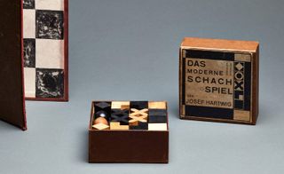 A chess set featuring the chess board on the left corner of the image, the chess open box showing the chess pieces and the cover of the chess box. Photographed against a light blue background