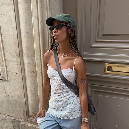 a woman wearing a greeb baseball cap, white lace camisole, and blue jeans