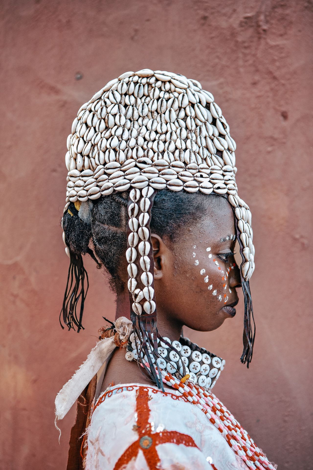 A woman adorned with a beautifully intricate shell headdress and traditional beaded attire stands against a textured earthy background