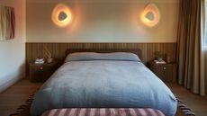 A bedroom with layered wall sconces, a wood paneled accent behind a large bed with blue bedding, matching wood side tables, a striped bench, and a patterned rug
