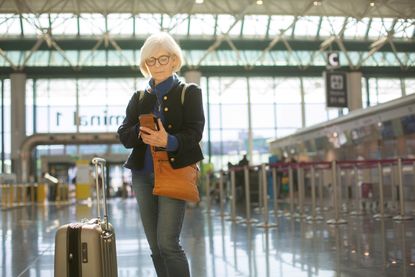A senior woman stands at the airport with her luggage while checking her phone. 