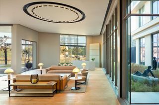 Elevate by Studio IAAD features face-on pair of brown sofas, and four round side tables with lamps on. Floor to ceiling windows.