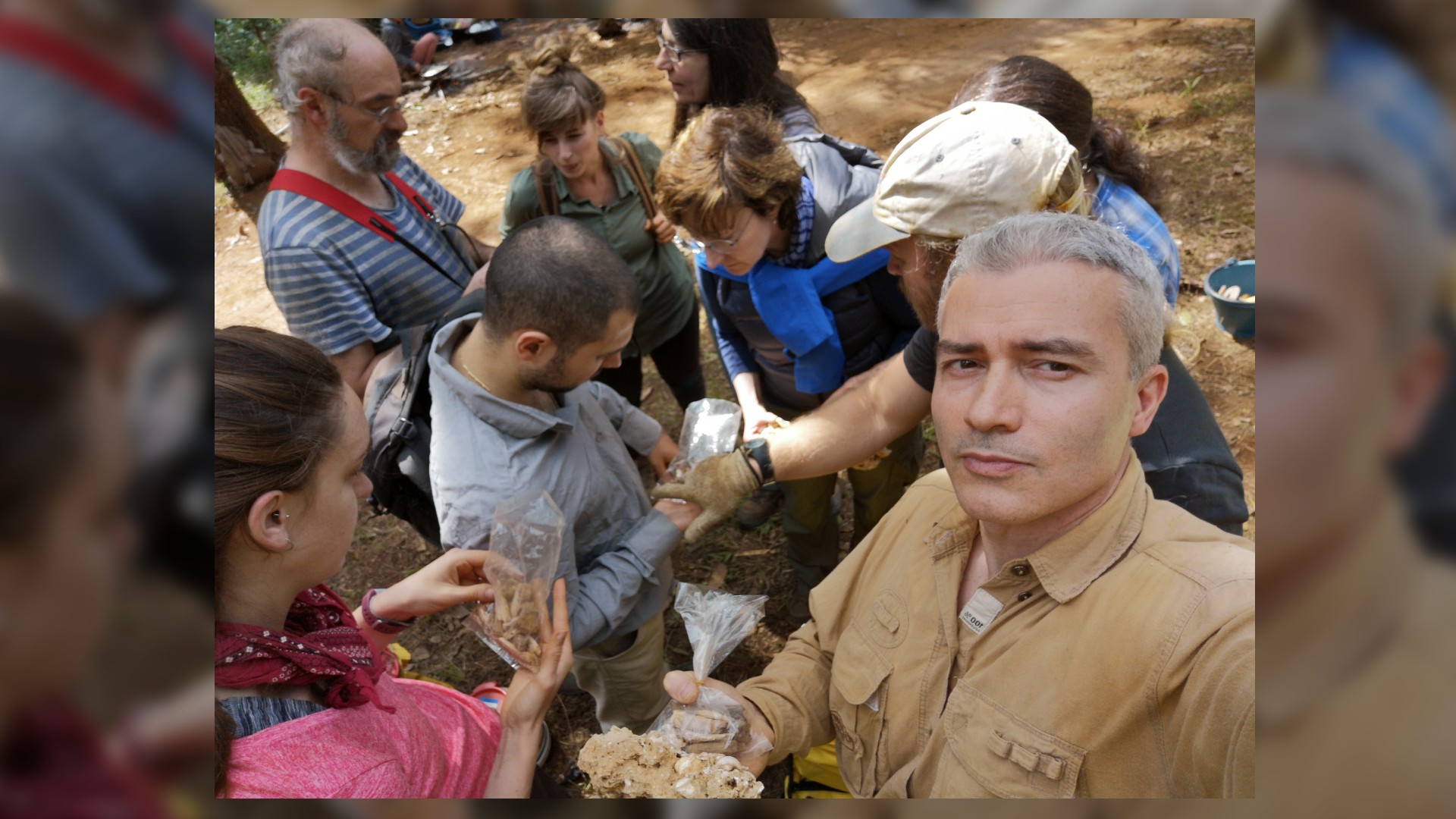 A group of nine researchers in a circle huddled over an interesting find. One man is facing the camera and proudly holding up a bagged artifact.