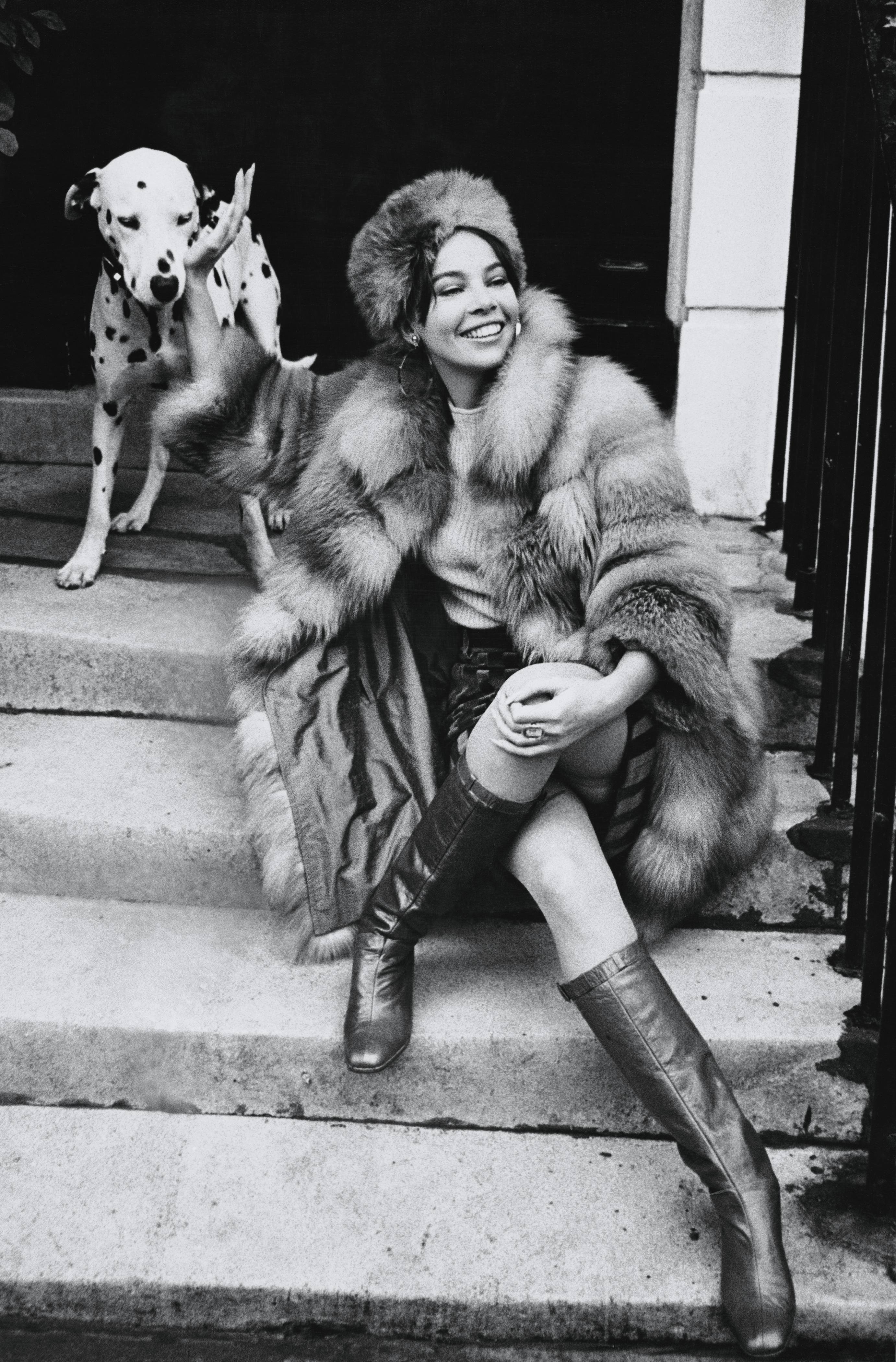 A black-and-white photograph of a smiling woman in a fur coat sitting on stone steps beside a dalmatian, which stands slightly behind her.