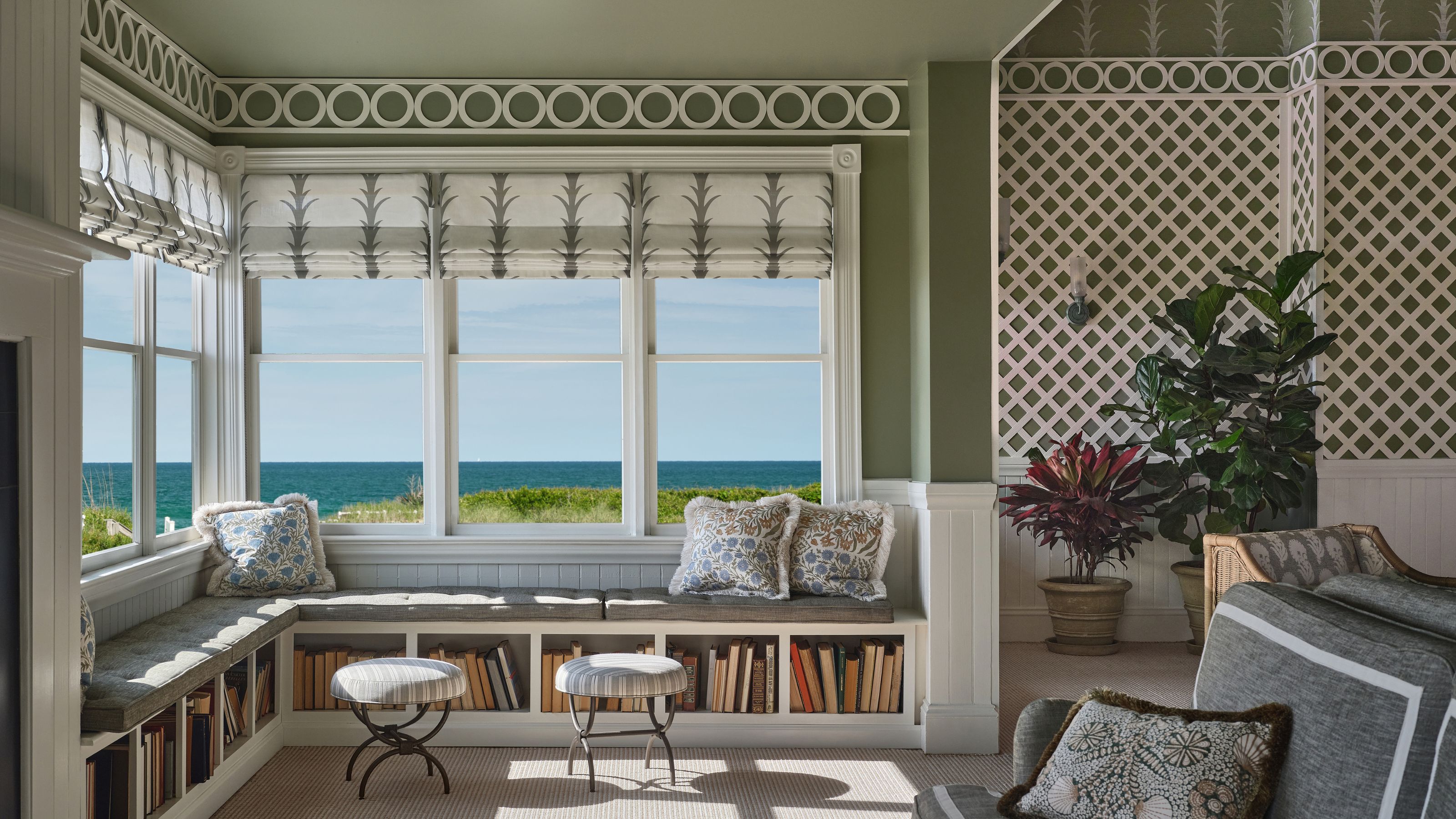 An L-shaped built-in window seat packed with books underneath, offering a panoramic view of the blue ocean, decorated with patterned throw pillows.