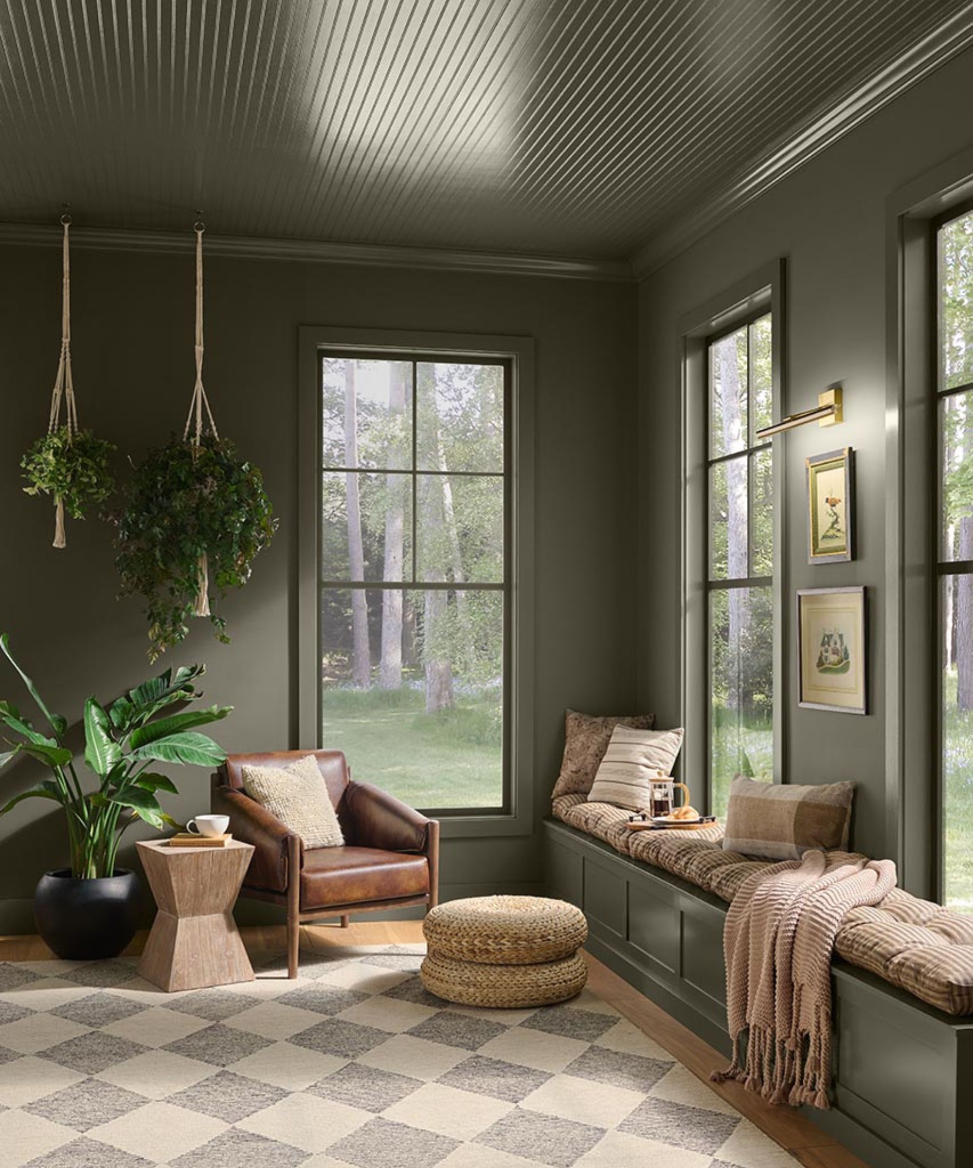 Sunroom painted olive green, three large long windows, a built-in bench seat, and a leather armchair and pouffe on top of a checkered rug.