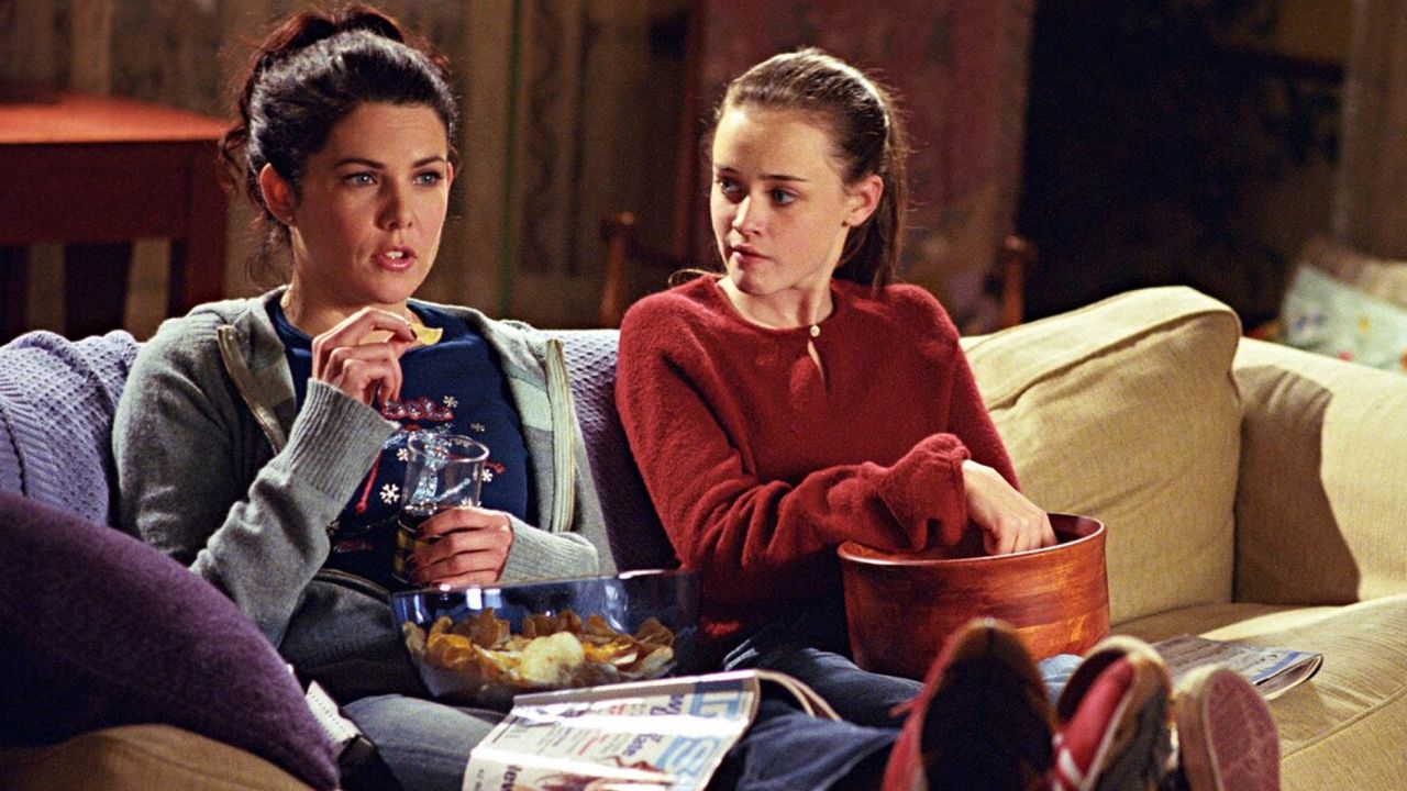 lauren graham as lorelai and alexis bledel as rory sitting on the couch eating snacks in gilmore girls