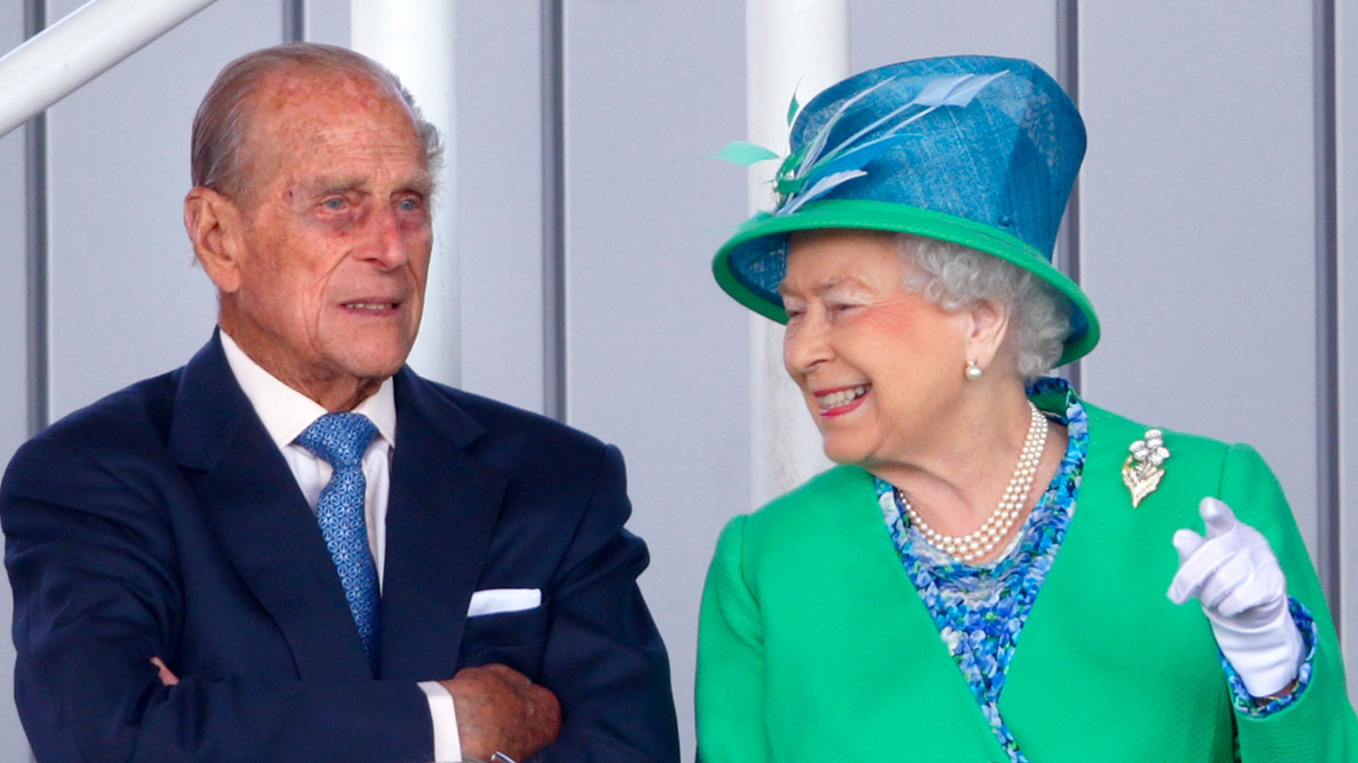 Queen Elizabeth wearing a green coat and blue hat smiling at Prince Philip who is folding his arms