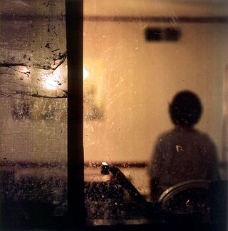 A photo taken from outside a kitchen window, through the glass and to a woman sitting at the kitchen table with her back to the camera