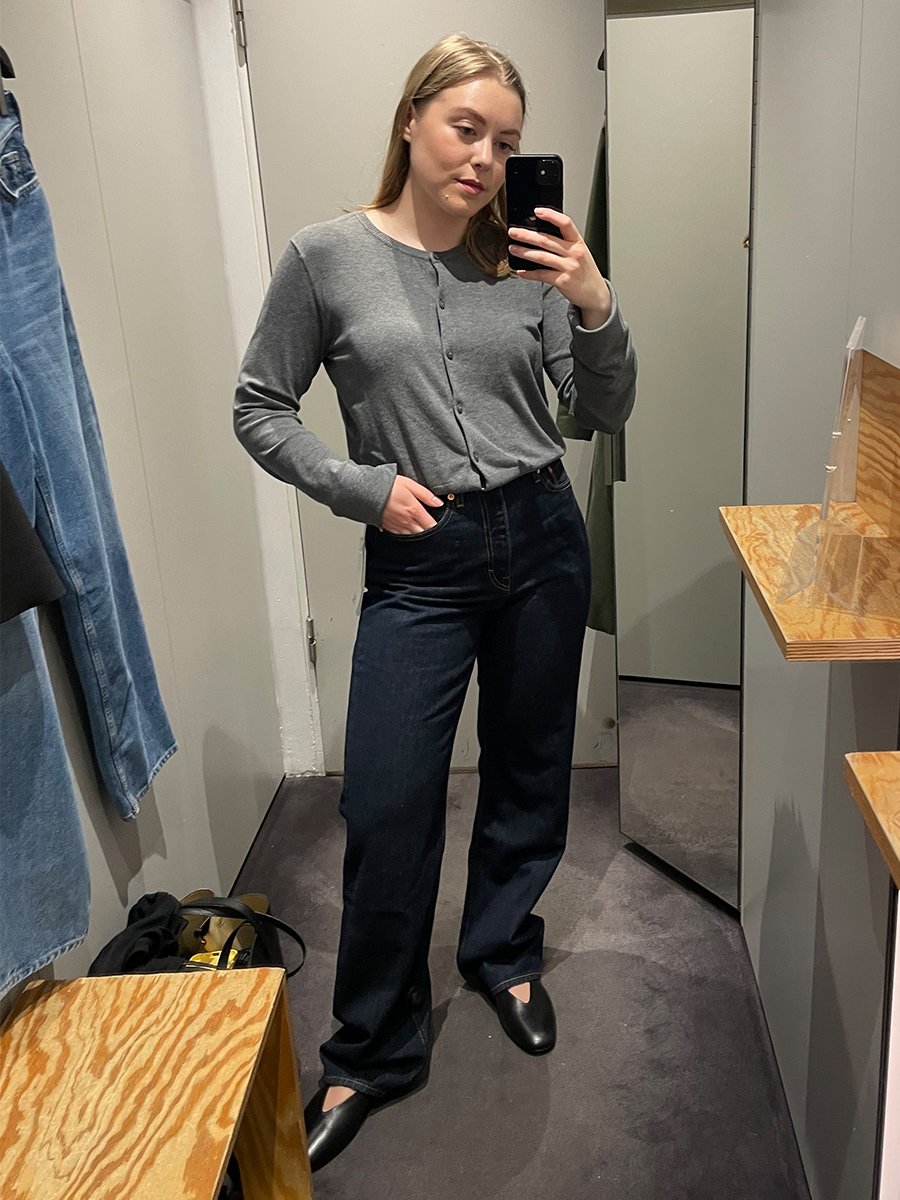 Woman in dressing room wears grey cardigan, dark blue jeans and mary janes