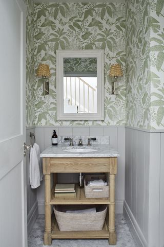 A light powder room with a wooden sink stand with open shelving, gray wall panels and green and white botanical wallpaper