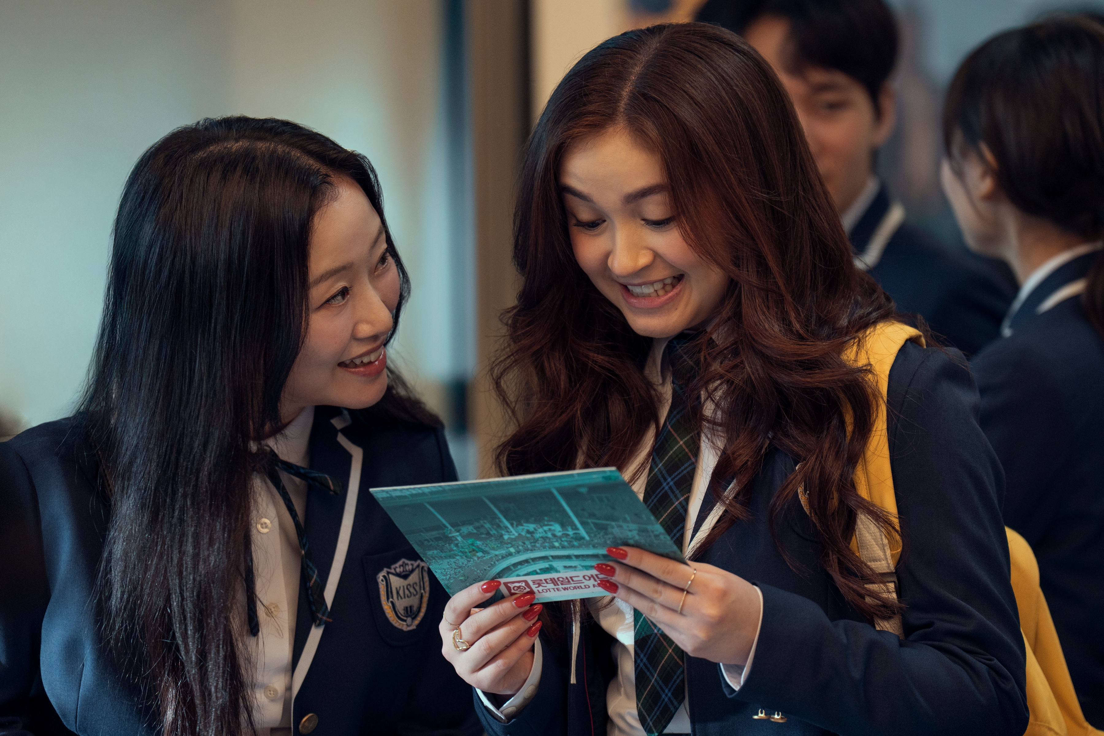 Gia Kim as Yuri Han and Anna Cathcart as Kitty Song Covey looking excitedly at a piece of paper while in school uniform and walking in the kiss hallway in xo kitty season 3