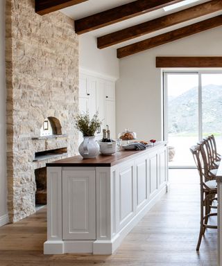 A French kitchen with a light brick wall with a bread and pizza oven, white walls, a dark brown countertop with a whitevase, tub with champagne, and a cake stand with a white panelled base, with dark brown beams above