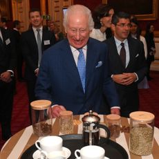 King Charles in a blue suit and tie standing and laughing in front of a table filled with cups, suacers and coffee supplies.