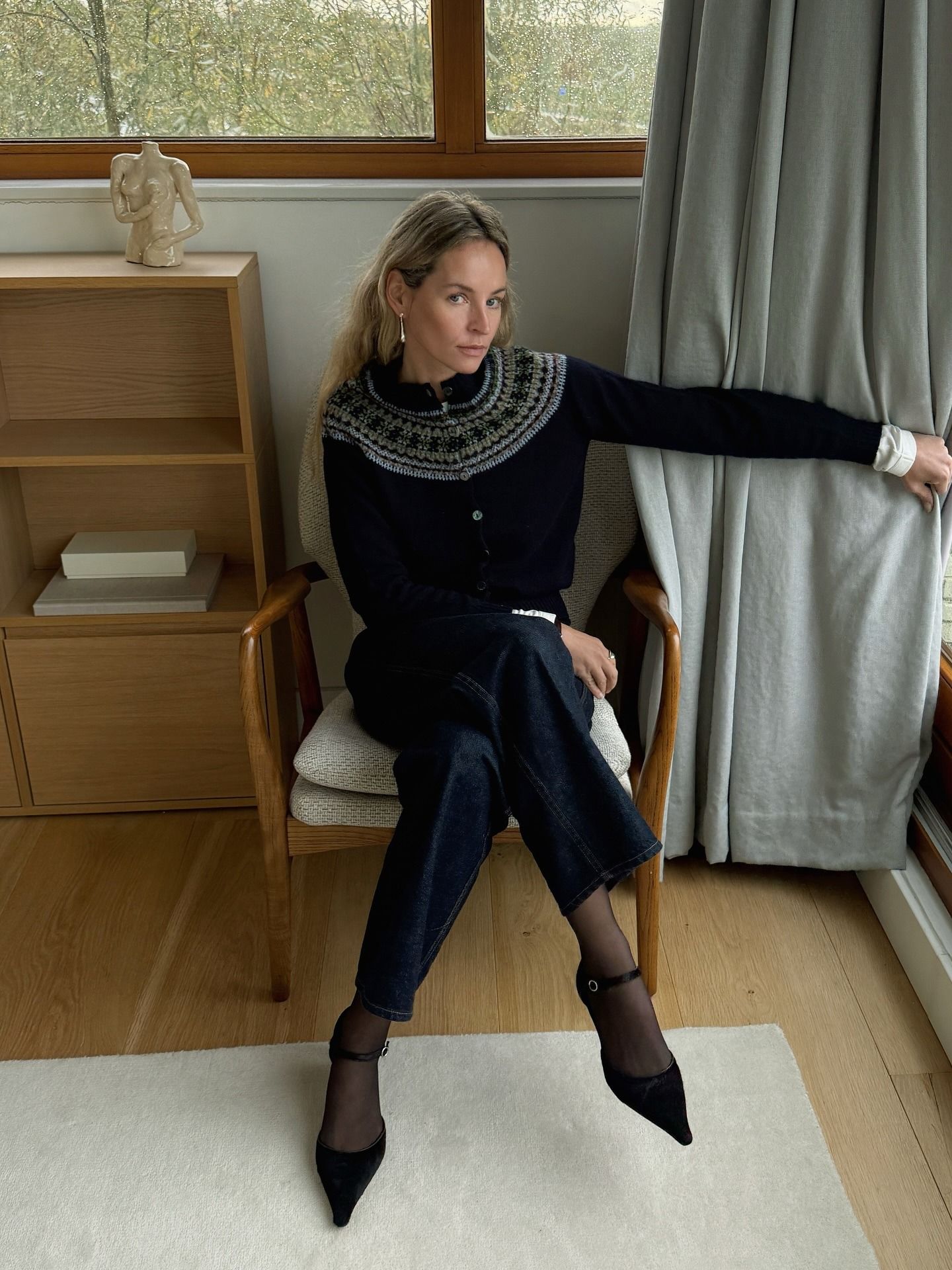 Influencer @anoukyve sits on a chair inside wearing a Fair Isle cardigan with dark wash jeans and pointed-toe heels.