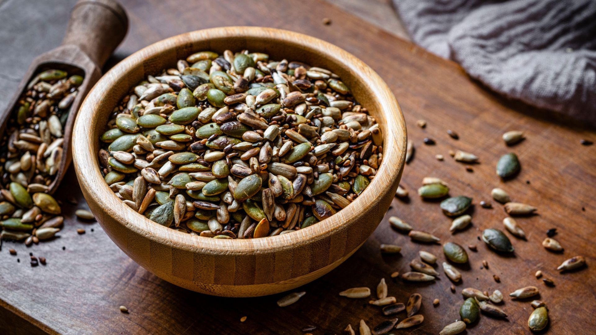 wooden bowl of toasted pumpkin seeds and some scattered around on wooden table