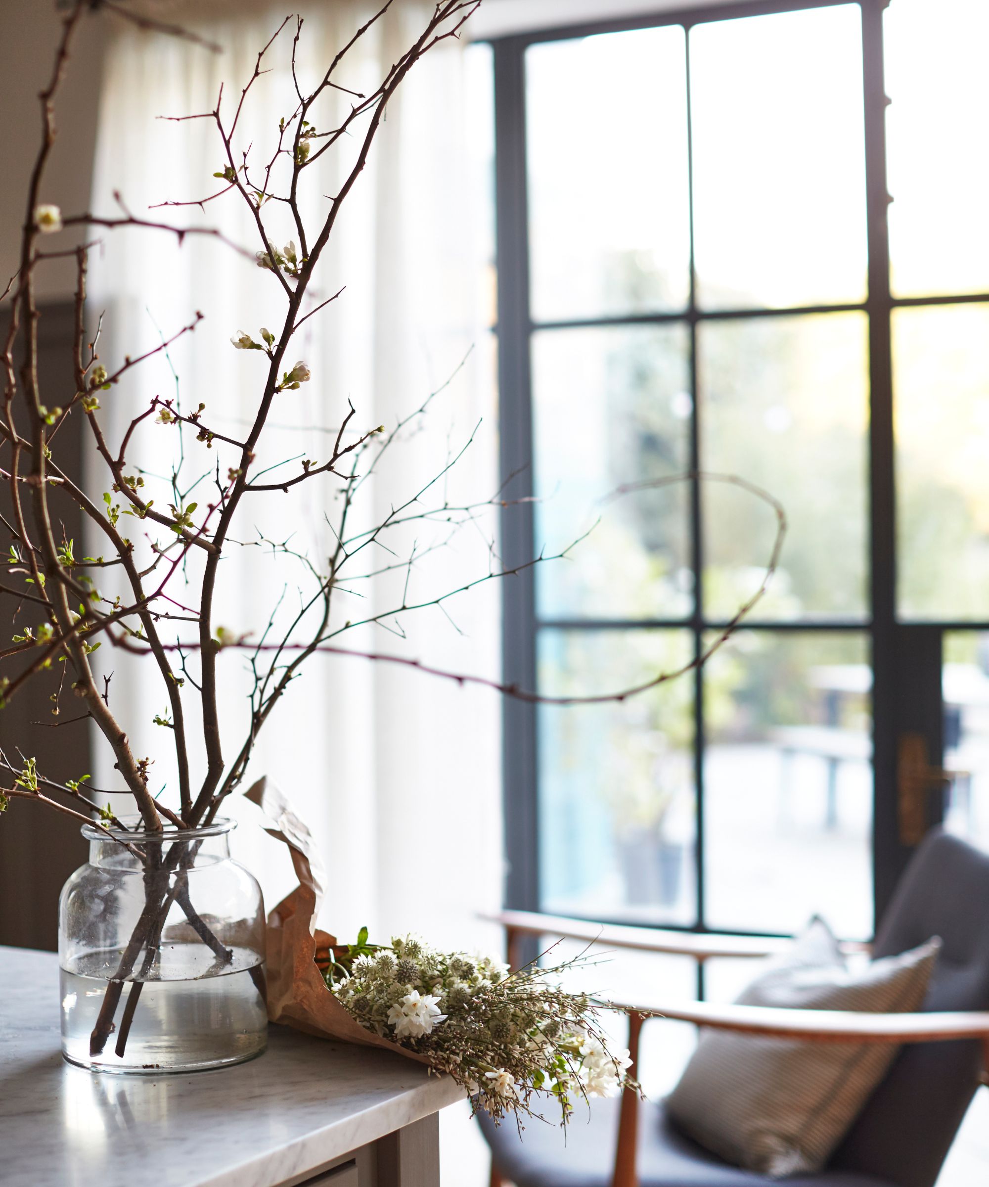 Blurred crittal windows with dried branches in a glass jar in the foreground, placed on a marble countertop next to some dried flowers. Behind is a chair with wooden arms and a grey cushion.