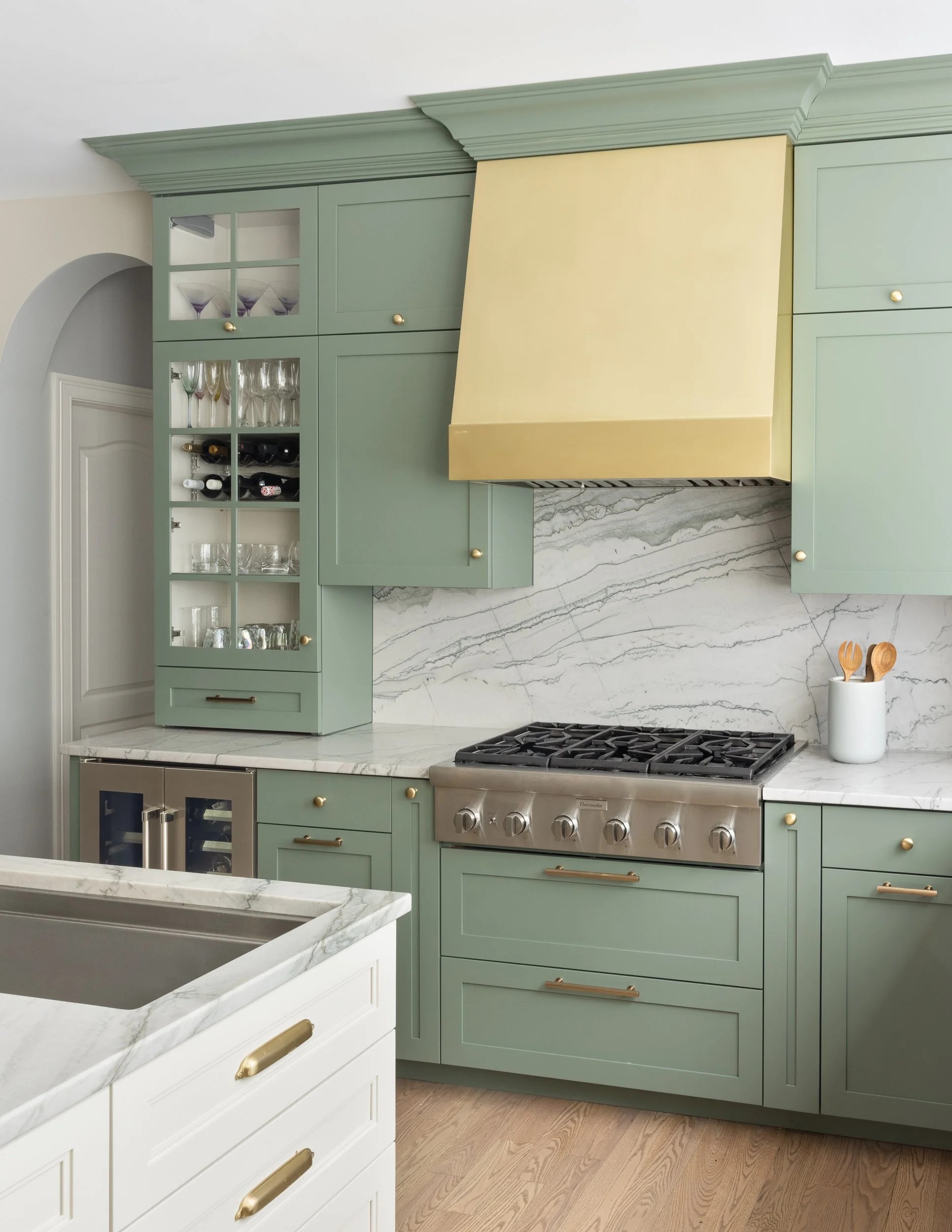 Image of a modern kitchen with sage green cabinetry and white marble countertops and backsplash. There is a gold cooker hood.